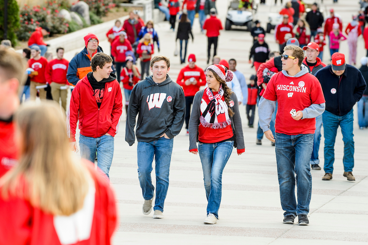 Crowd of people dressed in red Wisconsin apparel walk outdoors on a cool day.