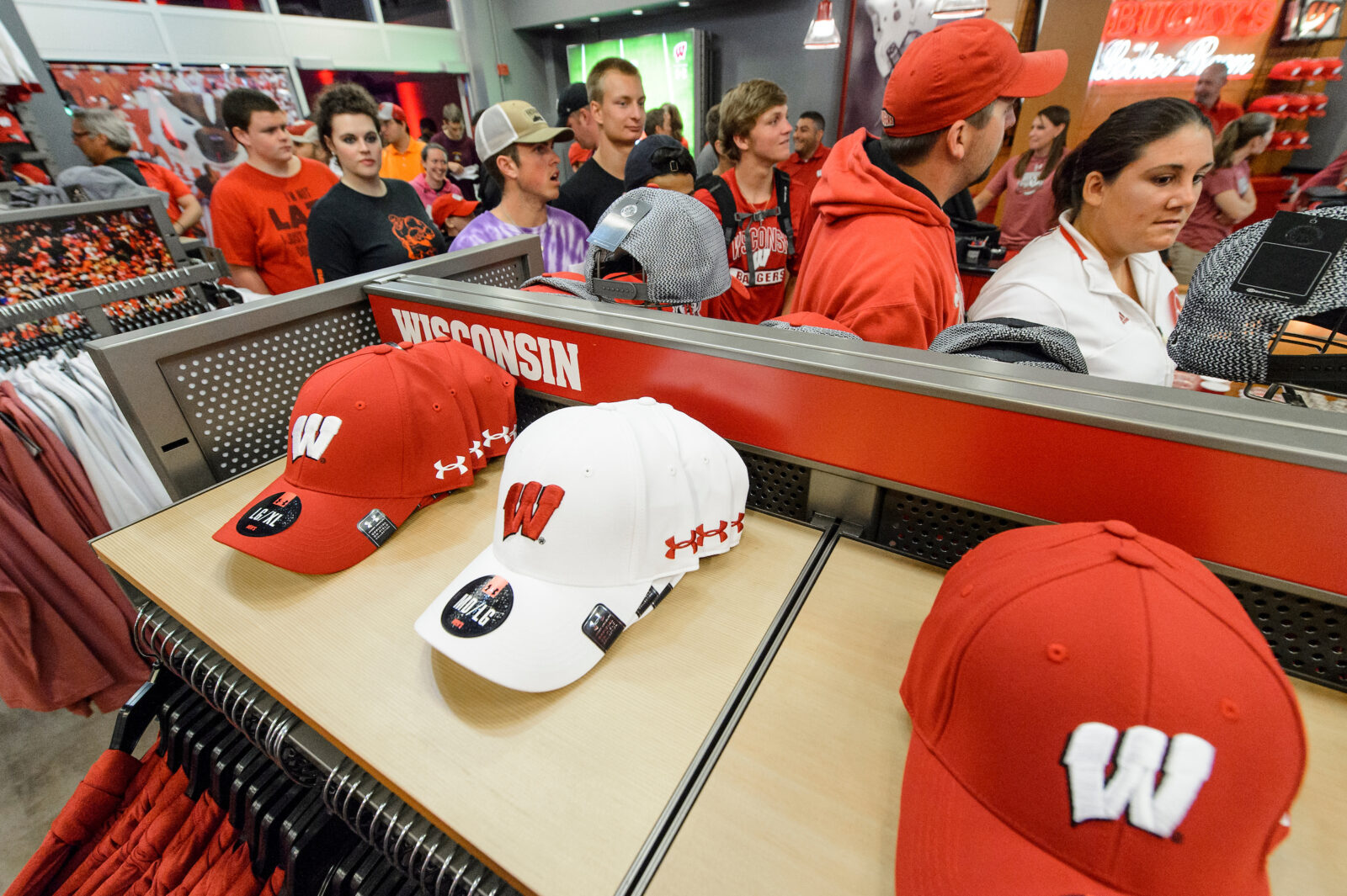 Red and white Motion W baseball caps displayed in the foreground, with shoppers in University of Wisconsin–Madison apparel browsing merchandise in background.